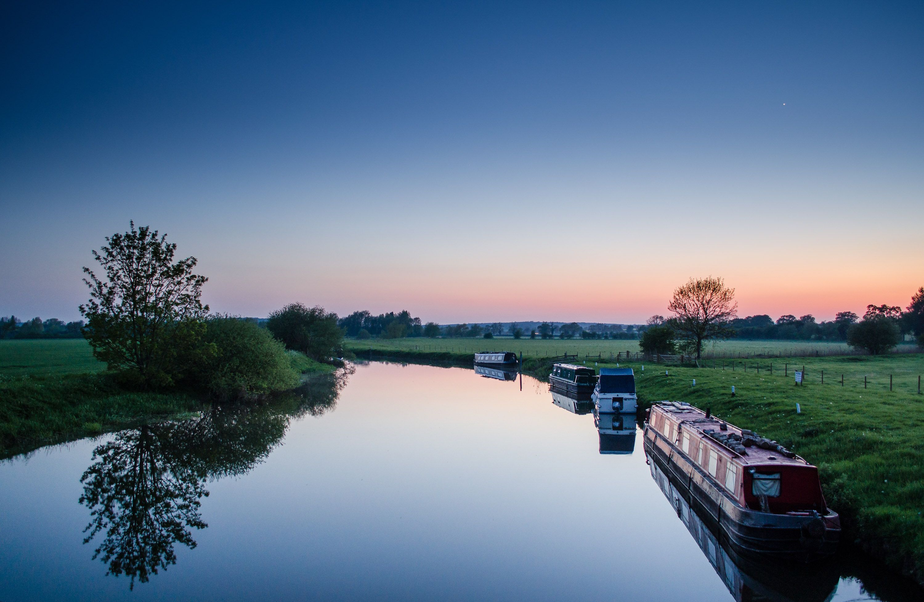 Several narrowboats moored along a calm canal at sunset, with trees and fields on the banks.